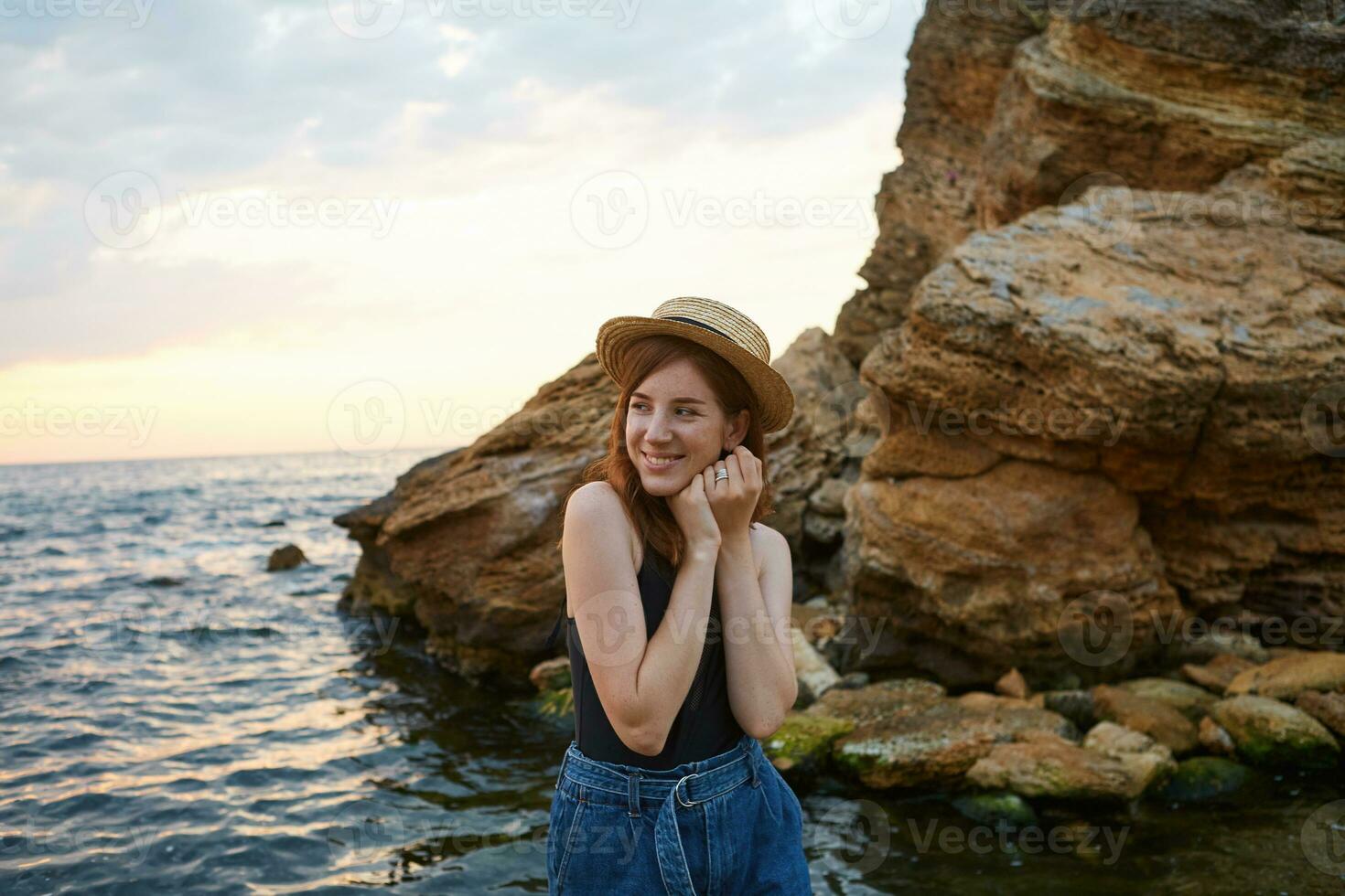 ung söt positiv ingefära flicka med fräknar bär i i hatt, brett ler och njut av de morgon- på de havet, utseende glad och Lycklig. foto