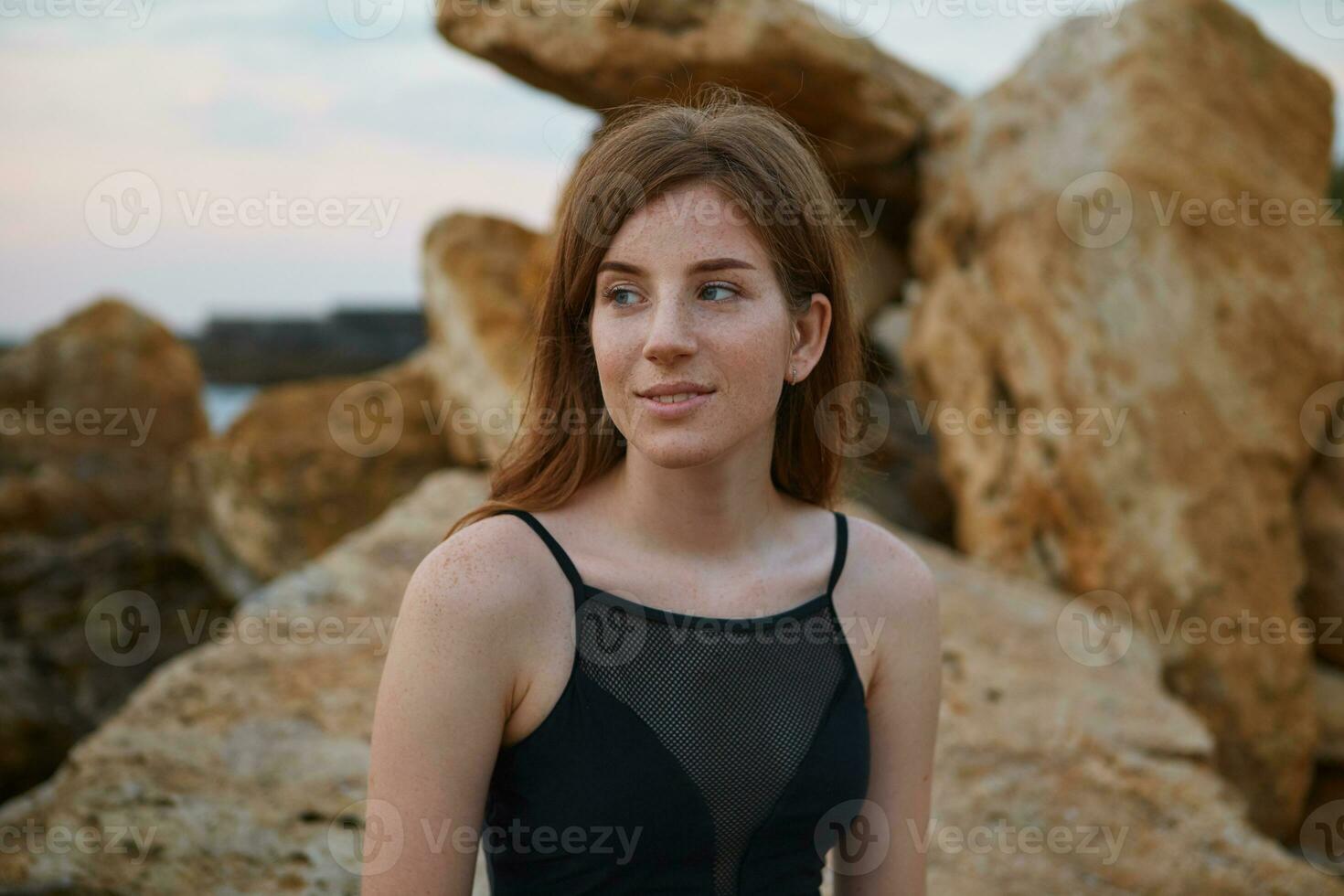 porträtt av ung leende ingefära söt kvinna med fräknar sitter på de strand, drömmande utseende bort, utseende positiv och Lycklig. foto