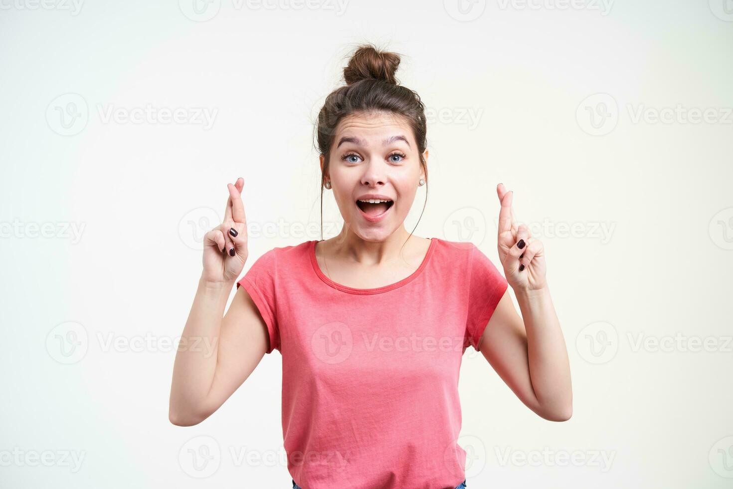 upprörd ung blåögd brun håriga kvinna med tillfällig frisyr korsning fingrar för Bra tur och ser upprymd på kamera, isolerat över vit bakgrund foto