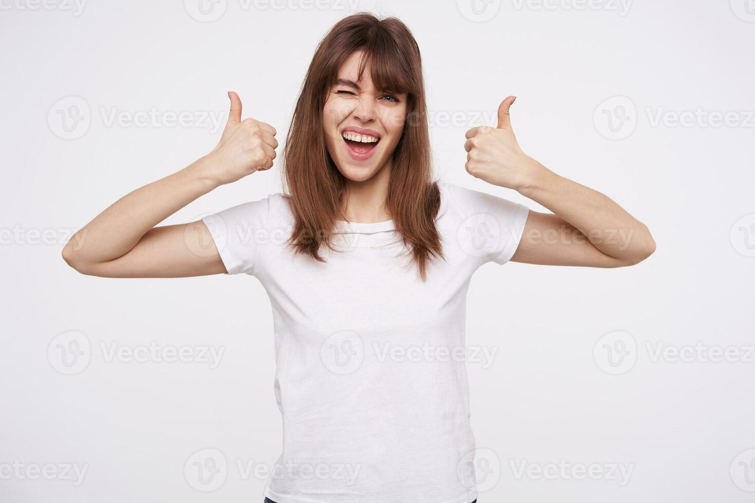 Lycklig ung skön brunett kvinna med tillfällig frisyr som visar Uppfostrad tummen medan ser glatt på kamera och ger blinkning, isolerat över vit bakåt foto