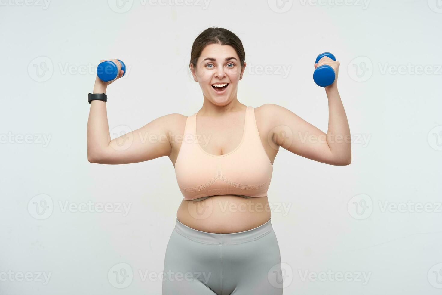 glad ung härlig brun håriga knubbig kvinna klädd i sportig ha på sig lyft hantlar och ser upprymd på kamera medan stående över vit bakgrund foto