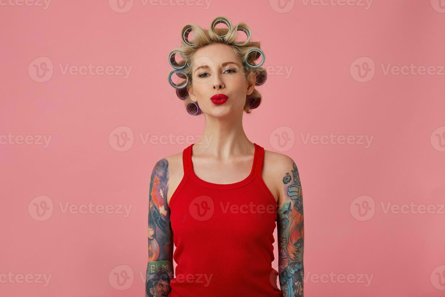 studio skott av charmig ung blond tatuerade lady framställning frisyr och hopfällbar henne röd mun i luft kyss medan stående över rosa bakgrund i röd skjorta med händer ner foto