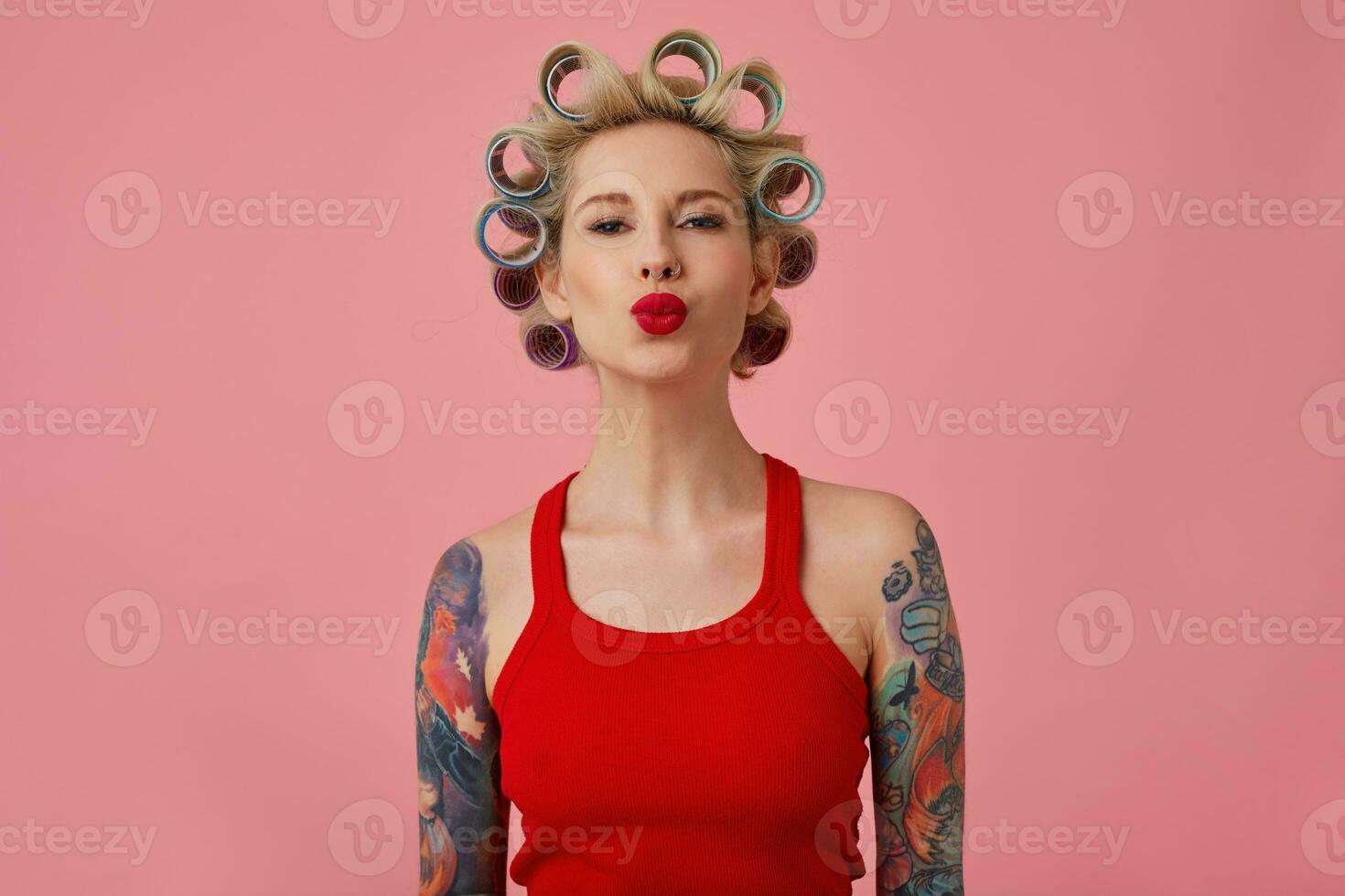 positiv härlig ung tatuerade kvinna curling henne blond hår på papiljotter medan framställning för kväll fest, hopfällbar henne röd mun i luft kyss medan ser på kamera, isolerat över rosa bakgrund foto