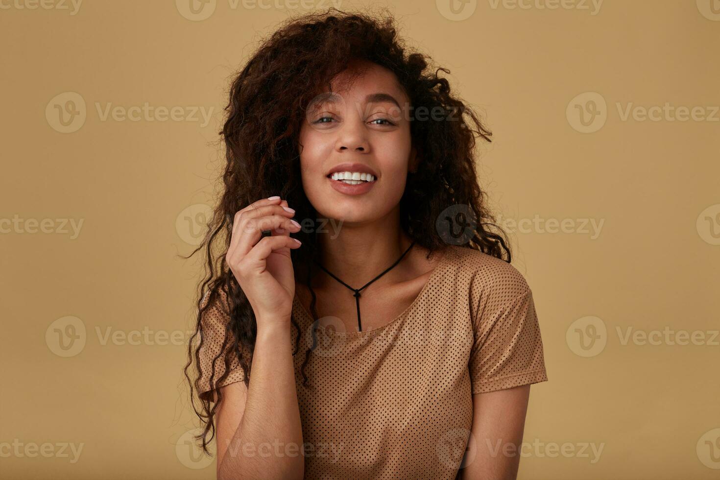 porträtt av ung glad lockigt mörk flådd brunett kvinna förvaring henne hand Uppfostrad medan ser positivt på kamera med trevlig leende, isolerat över beige bakgrund foto