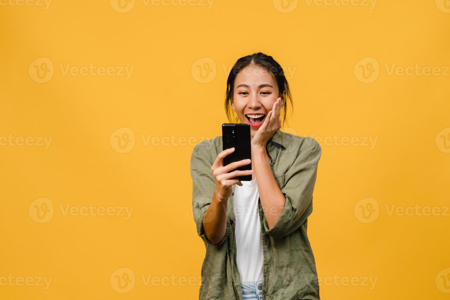 förvånad ung asiatisk dam med mobiltelefon med positivt uttryck, ler brett, klädd i vardagskläder och står isolerad på gul bakgrund. glad förtjusande glad kvinna jublar över framgång. foto