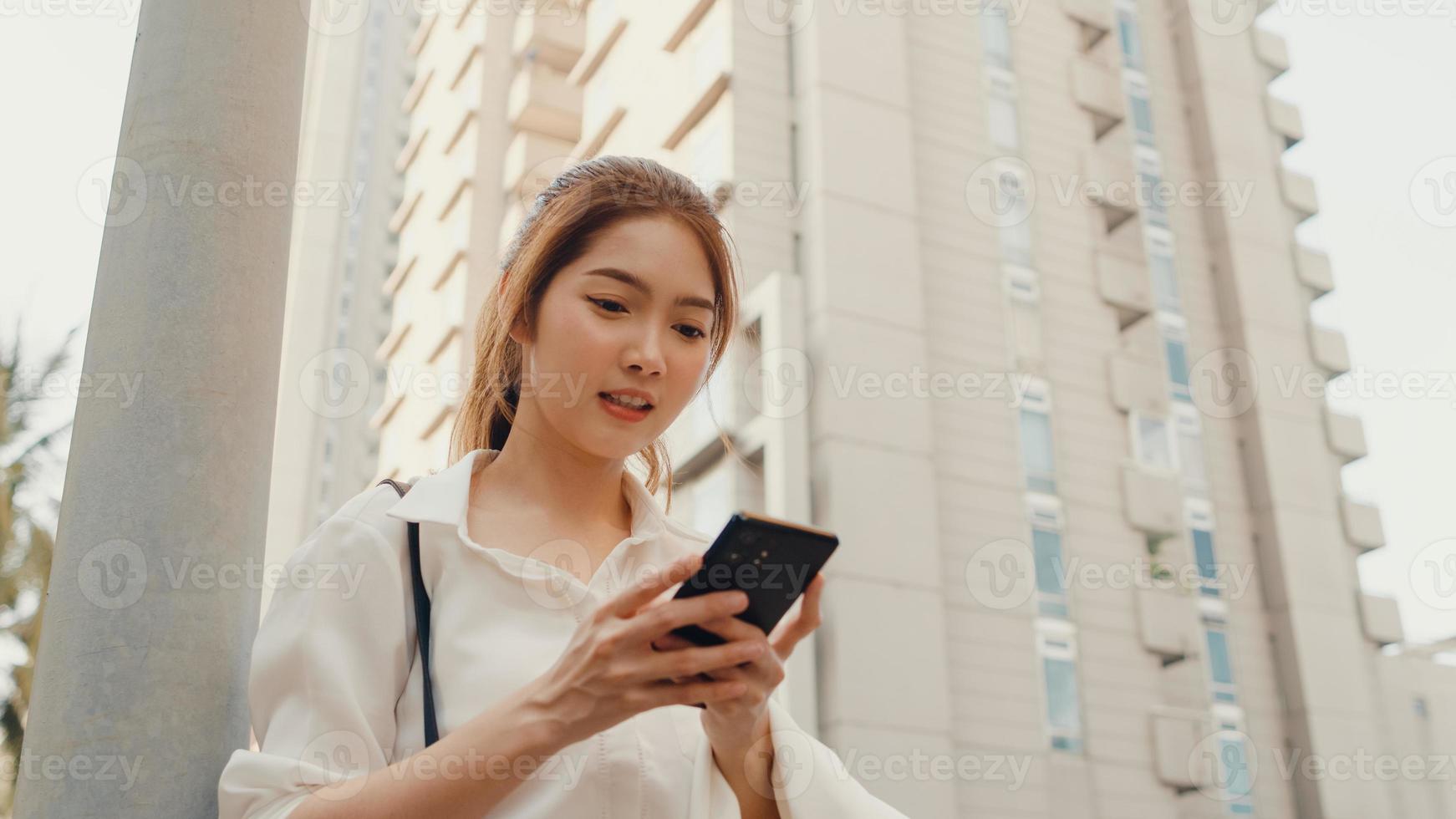 framgångsrik ung asiatisk affärskvinna i mode kontorkläder med smart telefon och skriver textmeddelande medan du går ensam utomhus i modern urban stad på morgonen. business on the go -koncept. foto