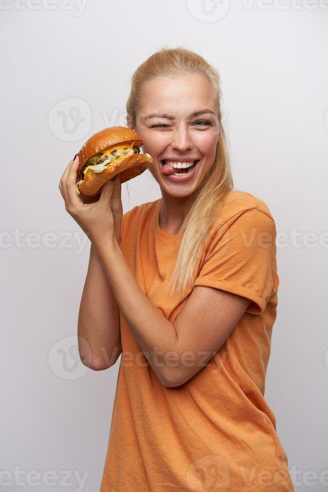 rolig skott av härlig ung blond kvinna med hästsvans frisyr ger blinkning till kamera och som visar henne tunga medan Framställ över vit bakgrund med färsk burger i henne händer foto