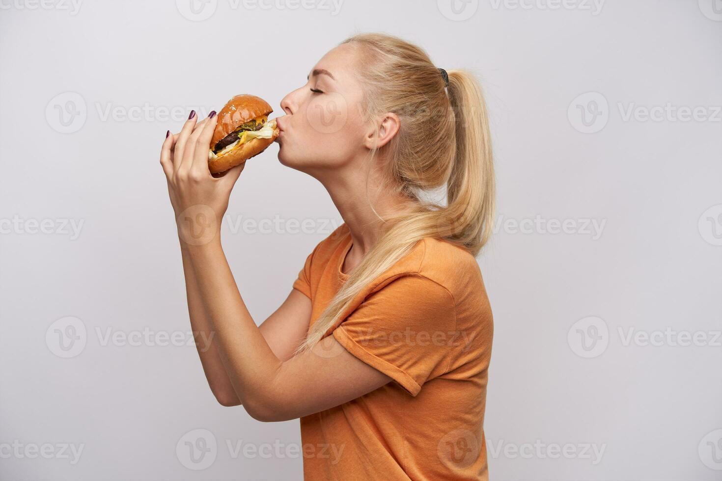 Bra ser ung blond kvinna med tillfällig frisyr innehav gott hamburgare i henne händer och kissing den med stängd ögon, bär orange t-shirt medan Framställ över vit bakgrund foto