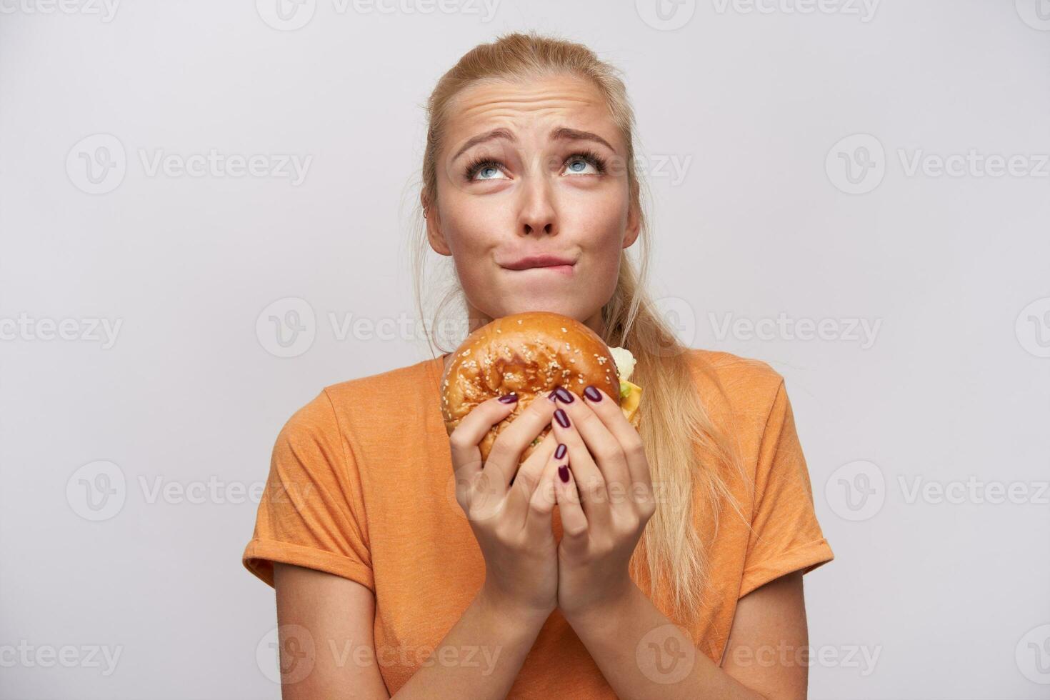 ung attraktiv lång håriga blond kvinna med hästsvans frisyr skrynklig panna och bitande underläpp medan ser ursäktande uppåt med skräp mat i händer, isolerat över vit bakgrund foto