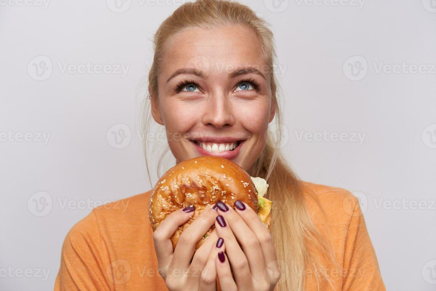 närbild av glad blåögd ung blond kvinna med stor gott hambuger ser lyckligt uppåt och leende allmänt, klädd i tillfällig kläder medan Framställ över vit bakgrund foto