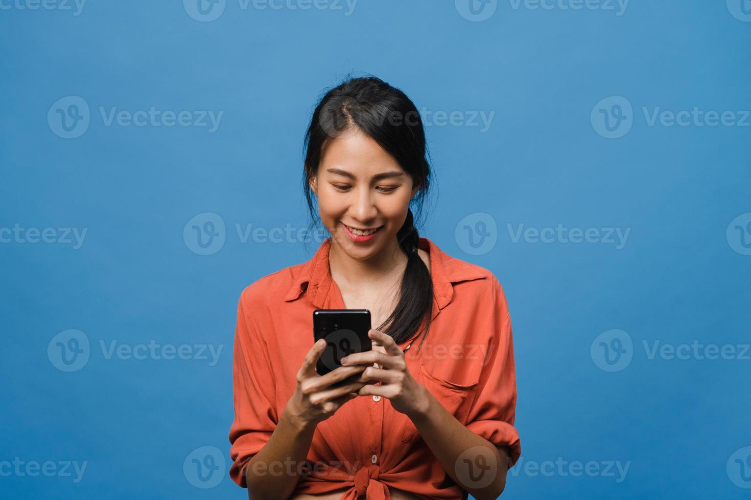 ung asiatisk dam som använder telefon med positivt uttryck, ler brett, klädd i vardagskläder som känner lycka och står isolerad på blå bakgrund. glad förtjusande glad kvinna jublar över framgång. foto
