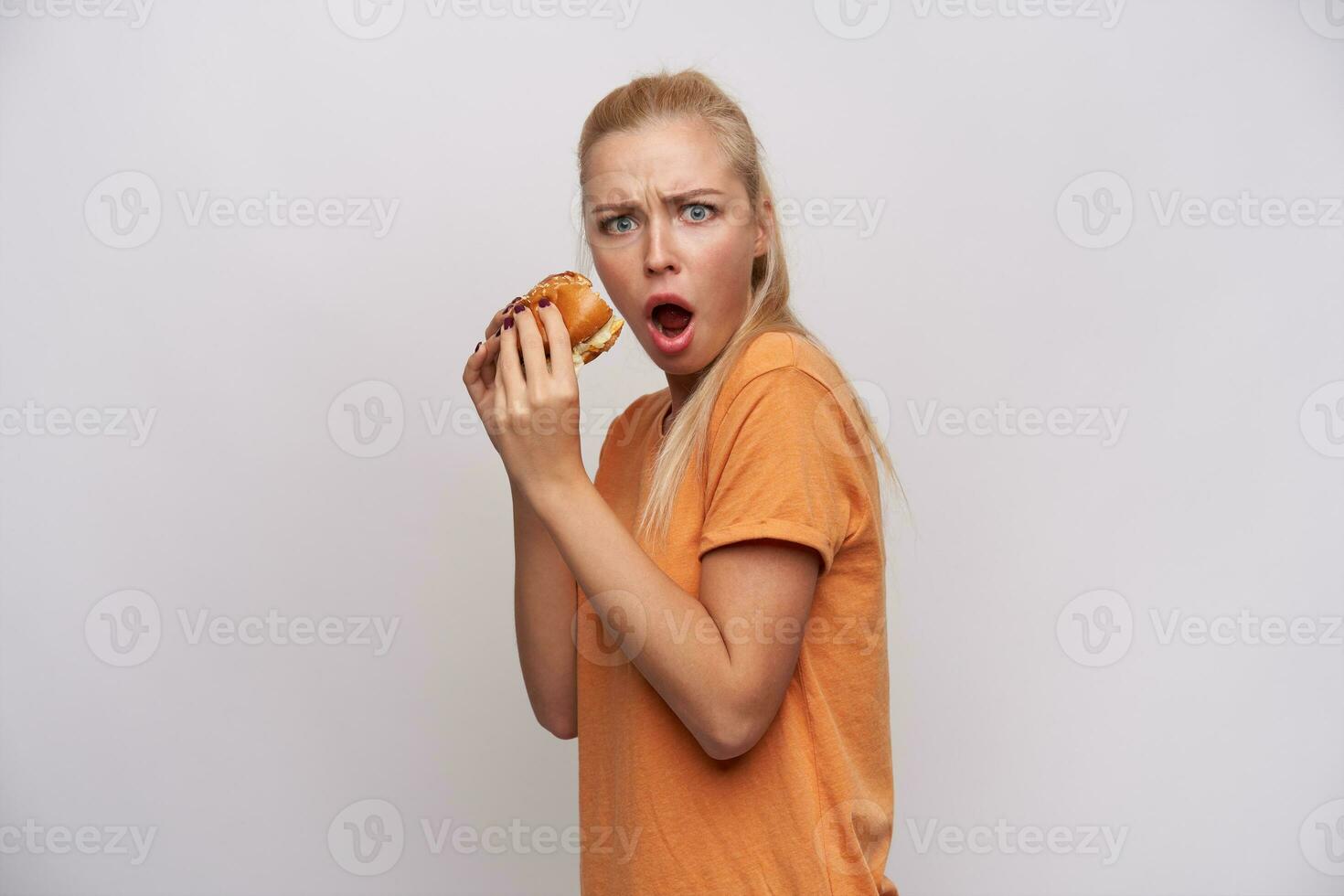 chockade ung attraktiv lång håriga blond kvinna med hästsvans frisyr innehav stor burger i henne händer och ser förvirrat på kamera, rynkar ögonbryn med öppnad mun över vit bakgrund foto