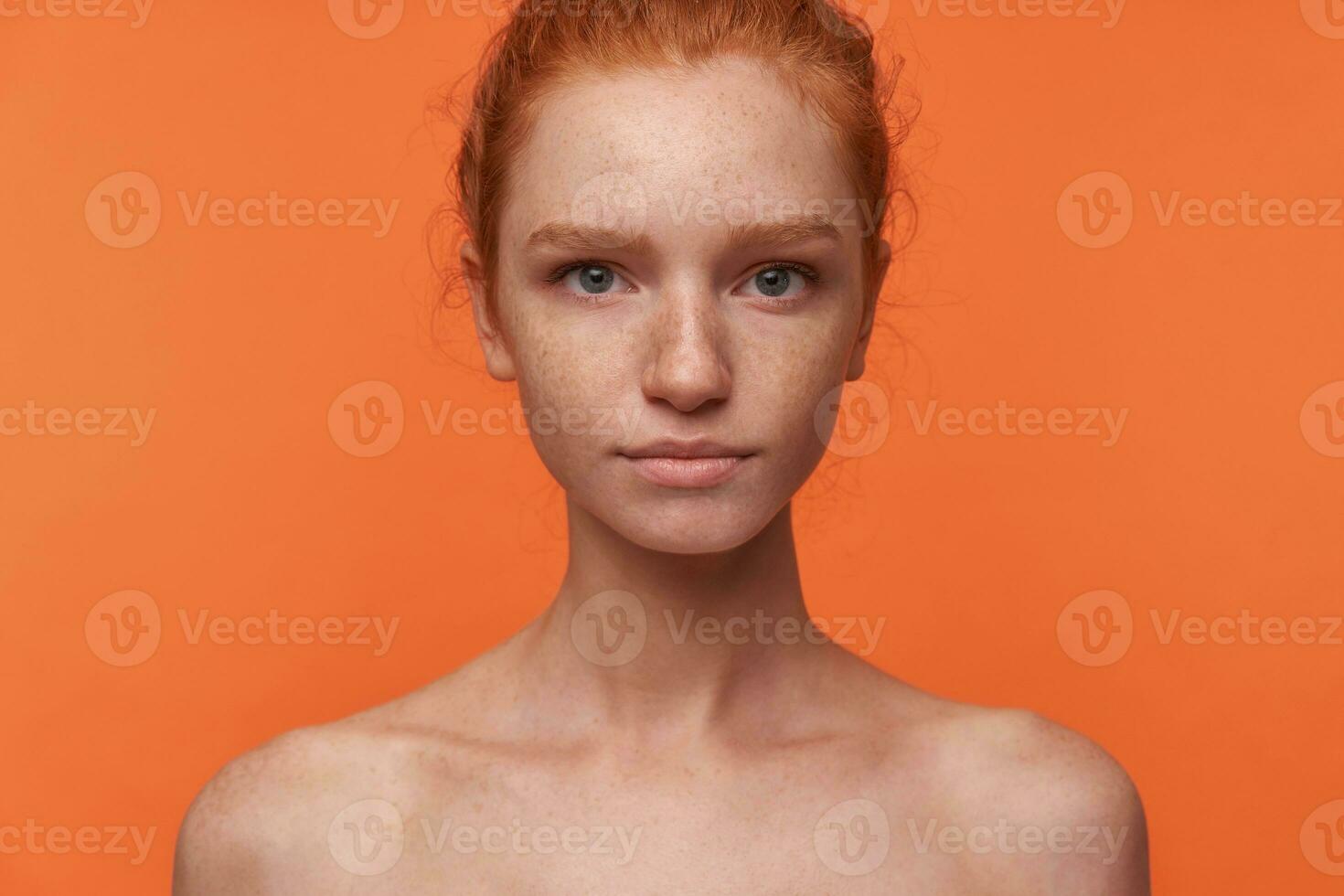 studio Foto av skön ung läshuvud kvinna med tillfällig frisyr stående över orange bakgrund utan kläder, ser på kamera med lugna ansikte