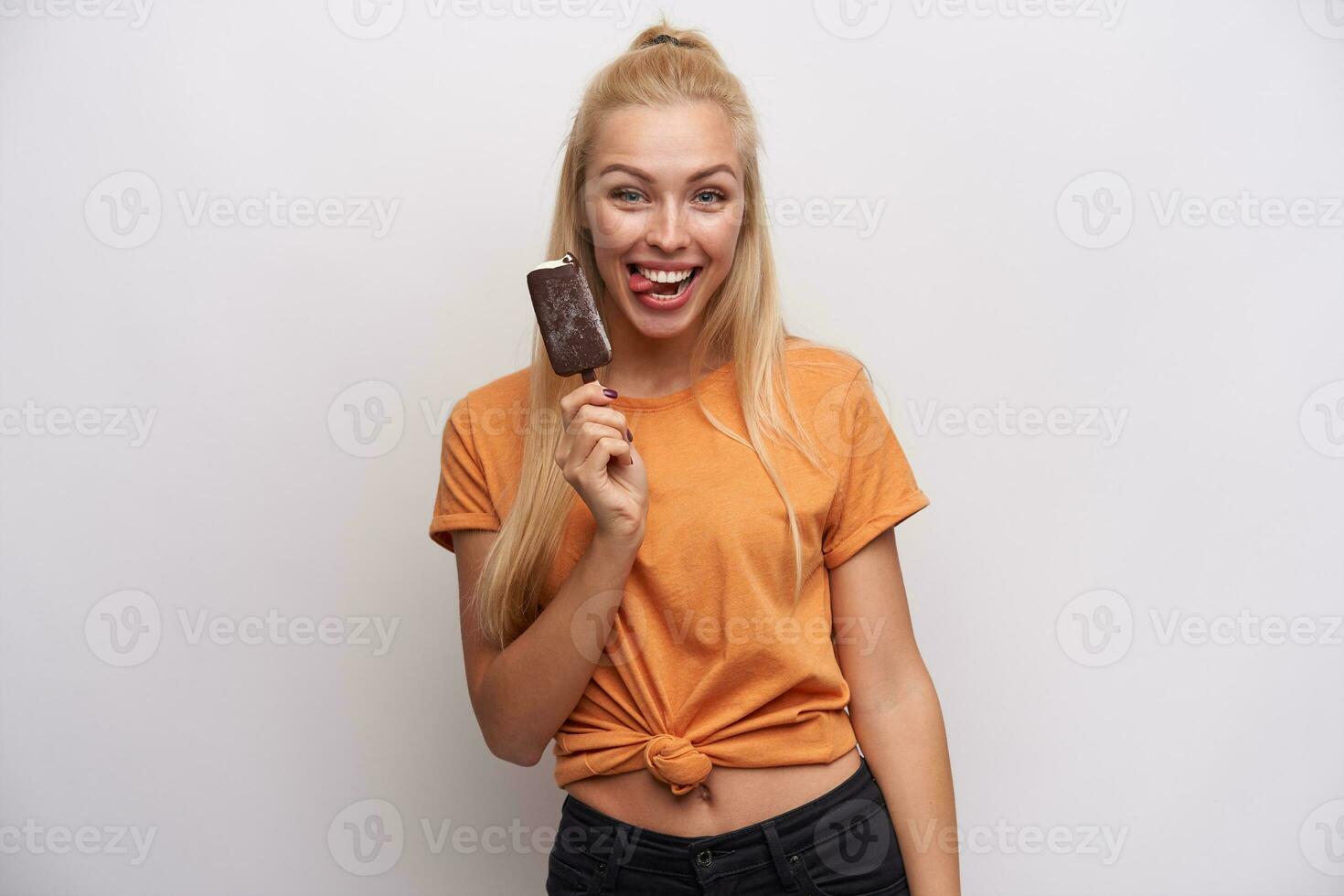rolig skott av glad Söt ung lång håriga blond kvinna i orange t-shirt som visar lyckligt tunga till kamera medan Framställ över vit bakgrund med glass foto