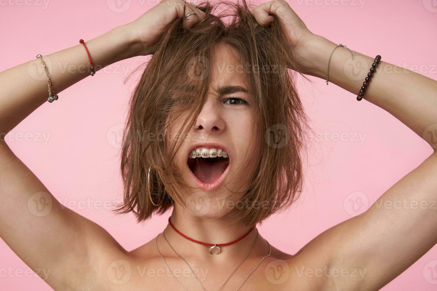 uttrycksfull ung kort håriga brunögd lady med kort frisyr ser upprymd på kamera med öppnad mun medan kläder henne huvud med Uppfostrad händer, isolerat över rosa bakgrund foto
