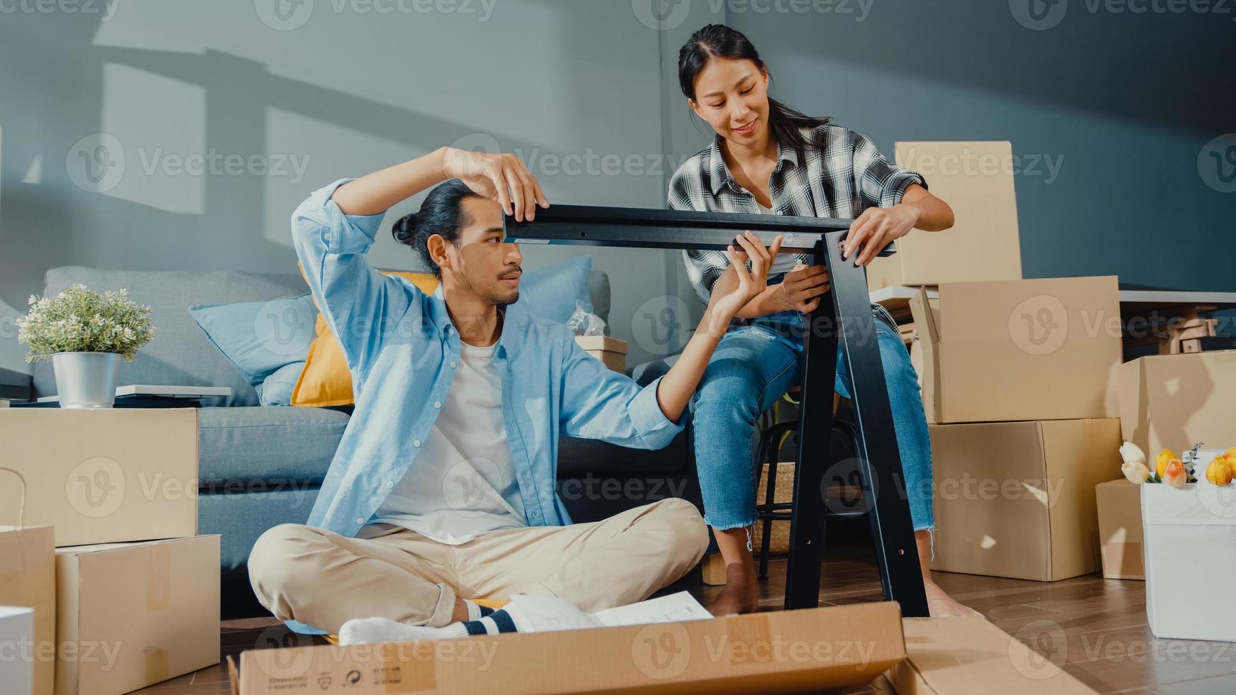 lyckliga asiatiska unga attraktiva par man och kvinna hjälper varandra att packa upp lådan och montera möbler dekorera husbygga bord med kartonglåda i vardagsrummet. unga gifta asiatiska flytta hem koncept. foto