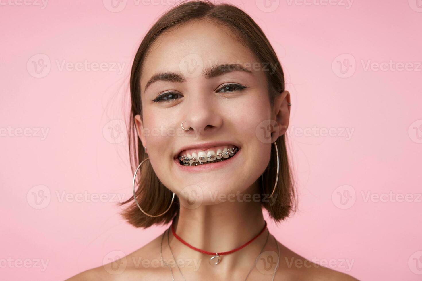studio skott av ung Söt glad kort håriga brunett kvinna med guppa frisyr varelse i trevlig humör och leende lyckligt till kamera, isolerat över rosa bakgrund foto
