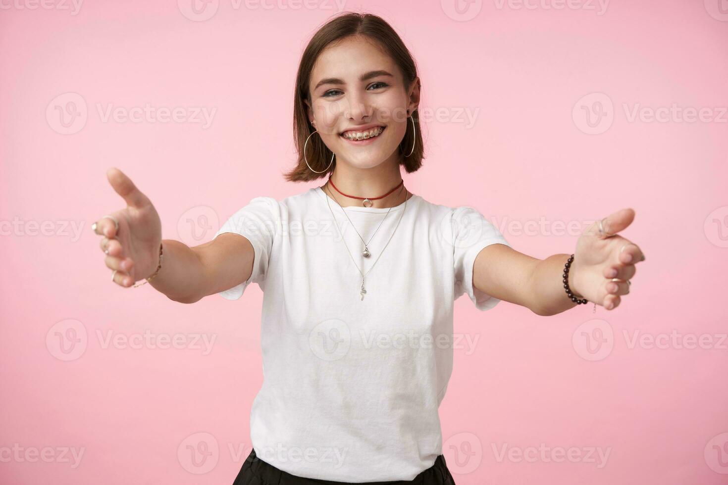 glad ung attraktiv kort håriga brunett lady med naturlig smink leende lyckligt medan stående över rosa bakgrund med bred vapen öppnad, gående till kram någon foto
