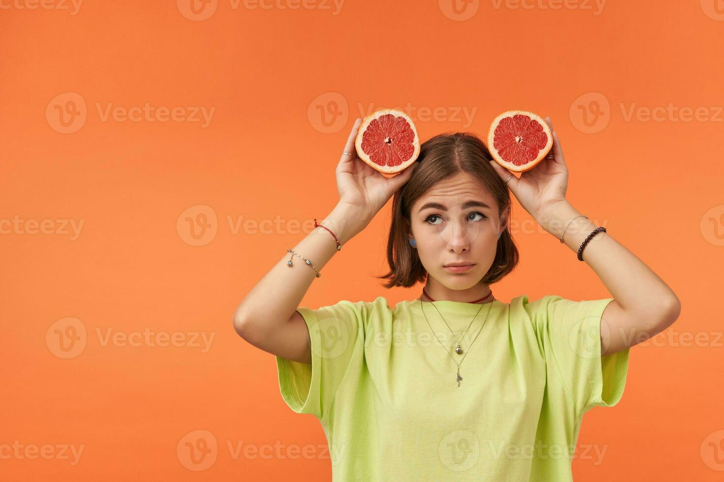 ung Söt kvinna med kort brunett hår, utseende ledsen, upprörd. innehav grapefrukt över henne huvud. stående över orange bakgrund. bär grön t-shirt, tandställning och armband foto
