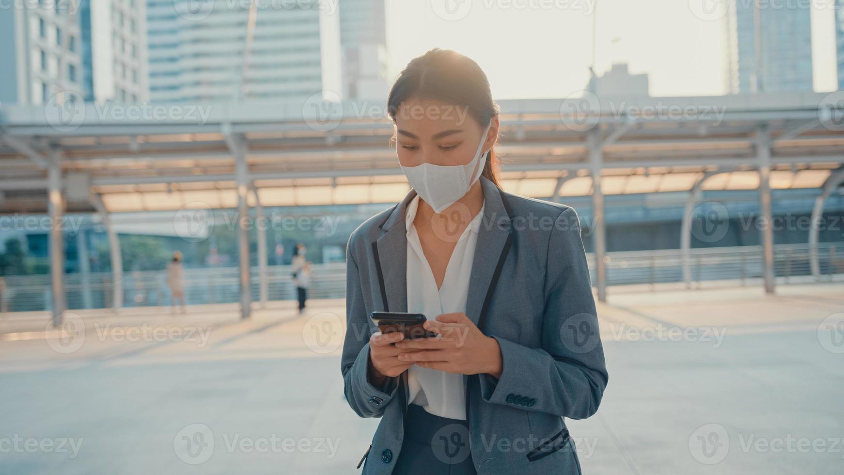 ung asiatisk affärskvinna i mode kontorkläder bär medicinsk ansiktsmask med telefon medan hon går ensam utomhus i stad. företag på gång, social distansering för att förhindra spridning av covid-19-konceptet. foto