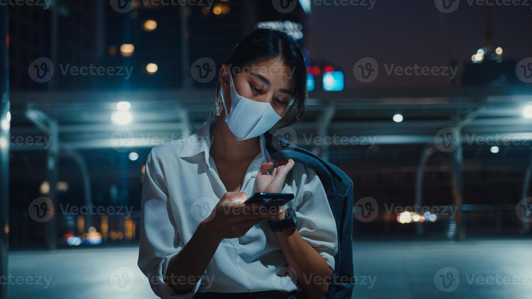 ung asiatisk affärskvinna i modekläder som bär ansiktsmask med hjälp av smarttelefon som skriver textmeddelande medan du står utomhus i stadens stad på natten. social distansering för att förhindra spridning av covid-19-konceptet. foto