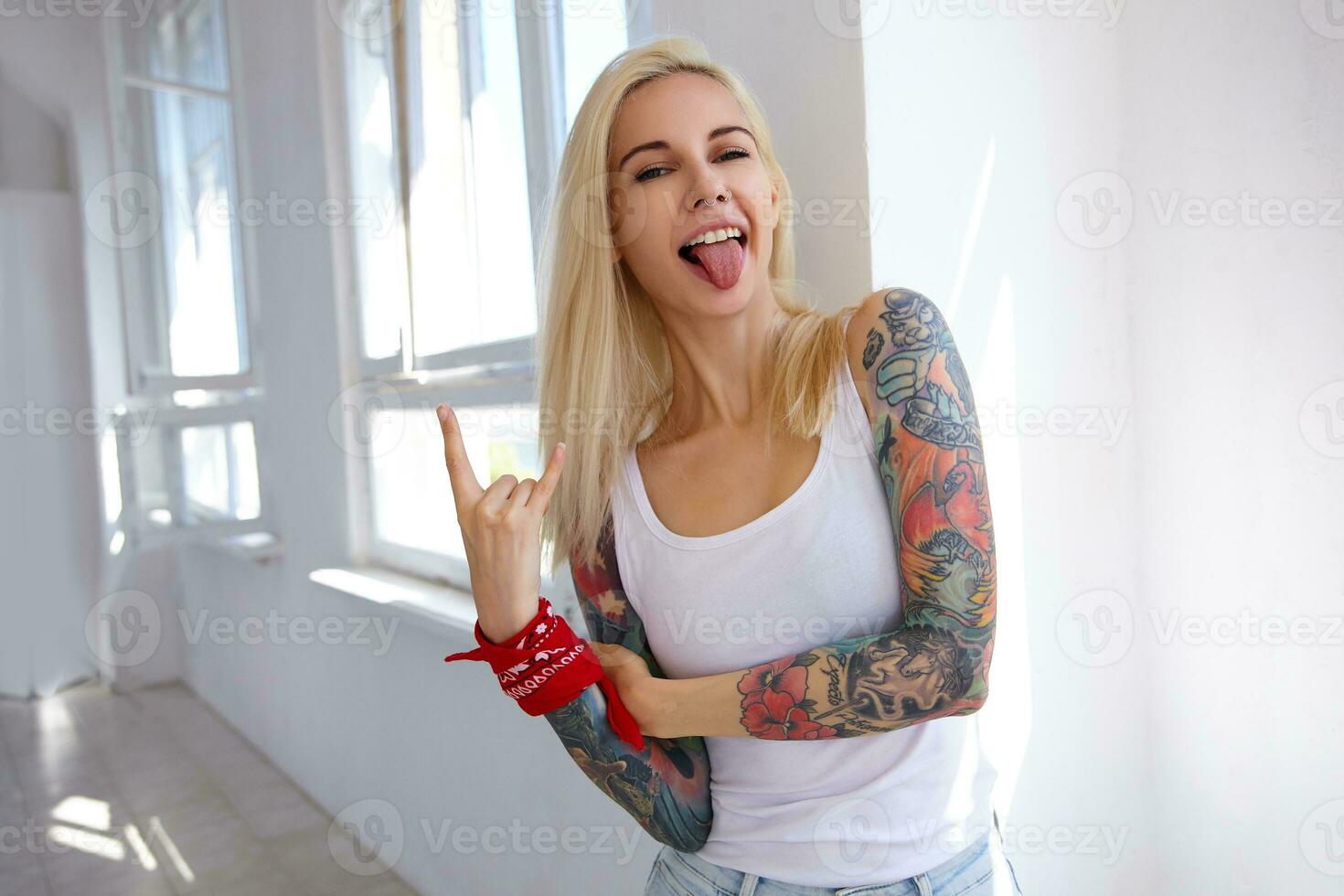 rolig skott av ung tatuerade Söt blond lady med naturlig smink som visar sten och rulla gest och fastnar glatt ut henne tunga, Framställ över ljus studio foto