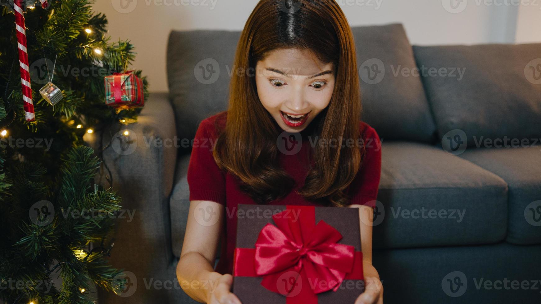 ung asiatisk kvinna som har roligt att öppna julklappslådan nära julgran dekorerad med prydnader i vardagsrummet hemma. god julkväll och gott nytt år. foto