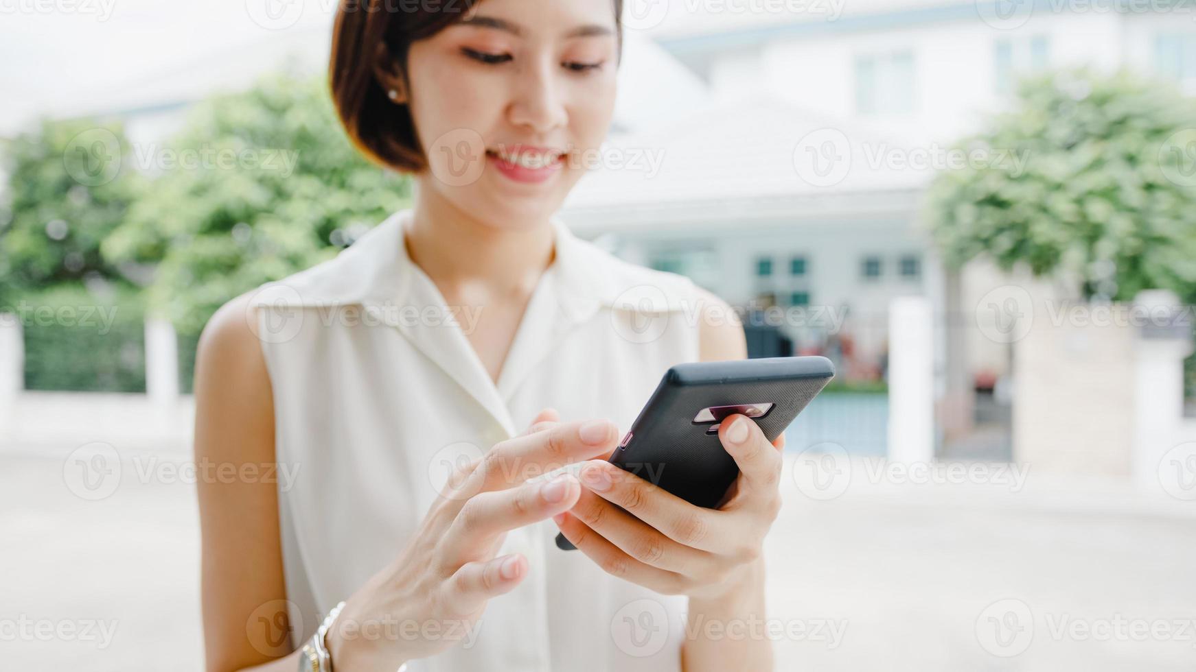 attraktiv ung asiatisk affärskvinna som använder mobiltelefon som kontrollerar sociala medier internet, chattar med vänner utanför på gatan i staden. livsstil ny normal efter coronavirus och social distansering. foto