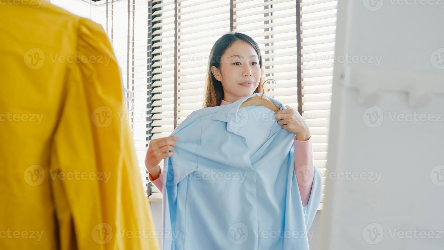 vacker attraktiv asiatisk dam som väljer kläder på klädhängare som ser sig själv i spegeln i sovrummet hemma. tjej tänk vad man ska ha på sig en casual skjorta. livsstil kvinnor slappna av hemma koncept. foto