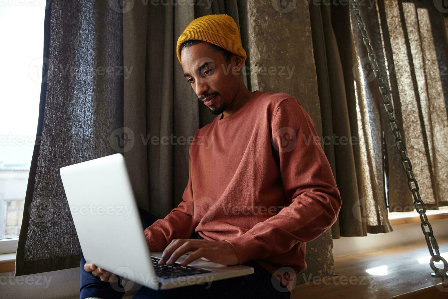 fundersam ung mörk flådd skäggig man i tillfällig kläder Sammanträde nära fönster på ljus dag och arbetssätt ut av kontor med bärbar dator, ser på skärm med koncentrerad ansikte foto