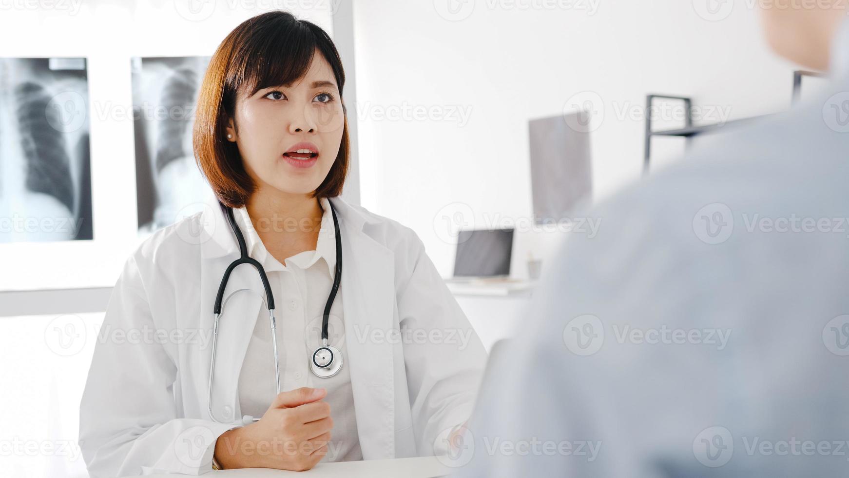 ung asiatisk kvinnlig läkare i vit medicinsk uniform som använder dator laptop levererar bra nyheter prata diskutera resultat eller symptom med manlig patient sitter vid skrivbordet på vårdkliniken eller sjukhuskontoret. foto