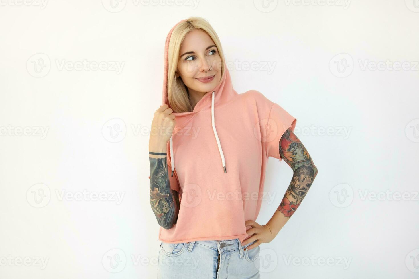 positiv ung skön tatuerade blond kvinna klädd i sportig kläder förvaring henne hand på midja medan ser gärna med trevlig leende, isolerat över vit bakgrund foto