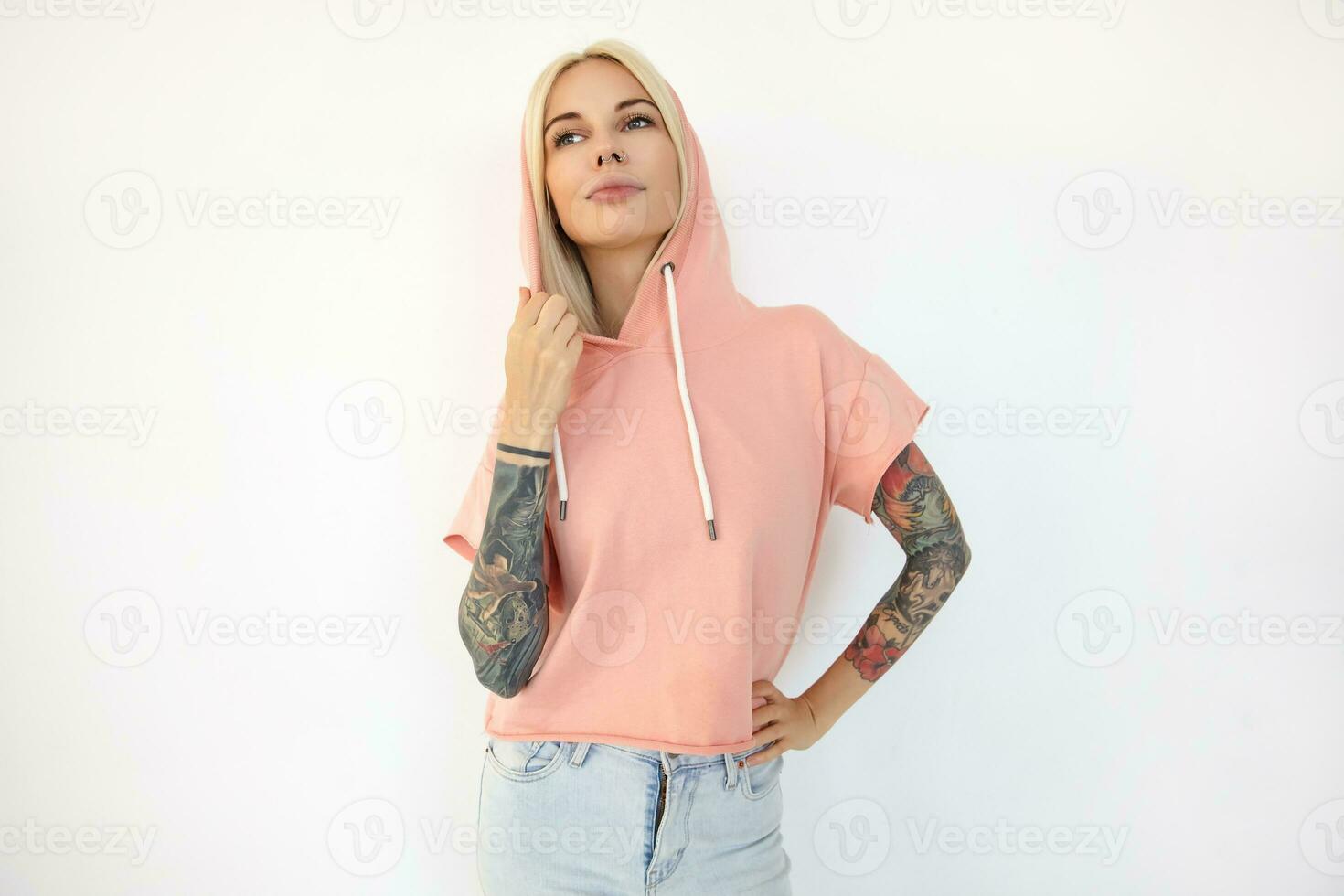 fundersam ung Söt tatuerade blond kvinna med näsa genomträngande bär rosa t-shirt med huva medan stående över vit bakgrund och ser eftertänksamt åt sidan foto