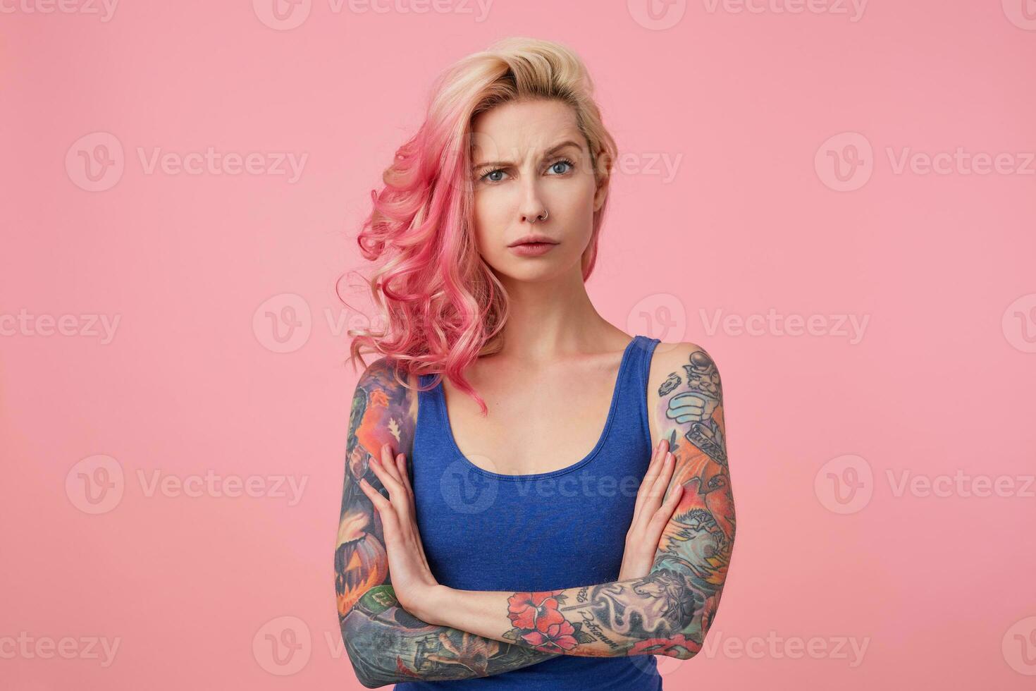 porträtt av ung missnöjd skönhet kvinna med rosa hår, rynkar pannan och står med korsade vapen över rosa bakgrund, utseende ledsen, bär en blå skjorta. människor och känsla begrepp. foto