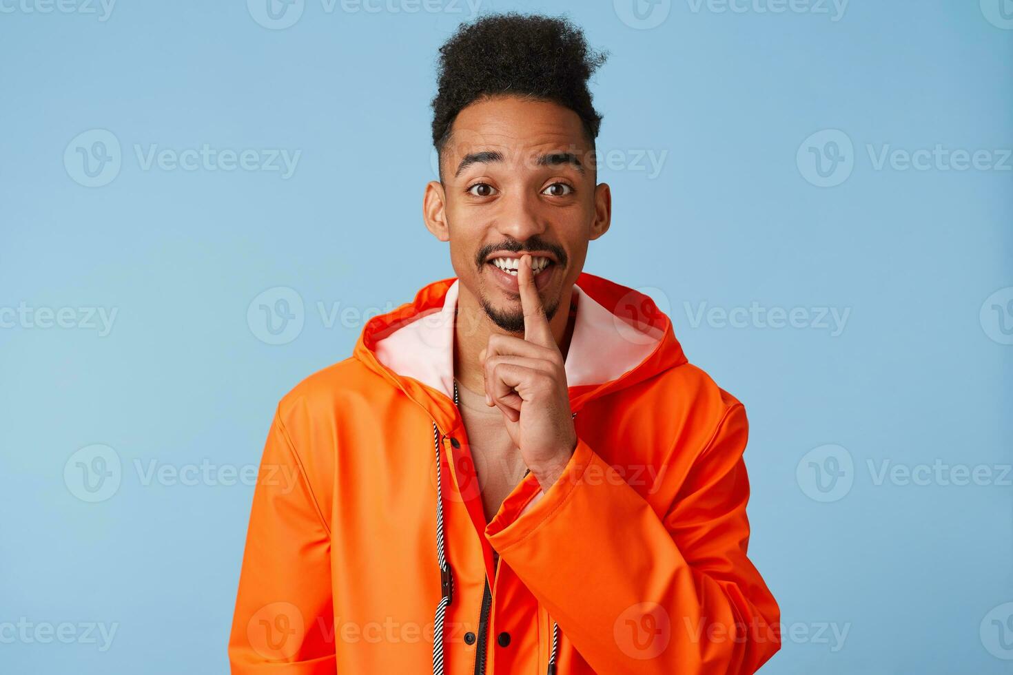 hemlighet ung afrikansk amerikan mörk flådd pojke bär i orange regn täcka har mystisk uttryck, finputsning mun med främre del finger, ler brett och står över blå bakgrund. foto