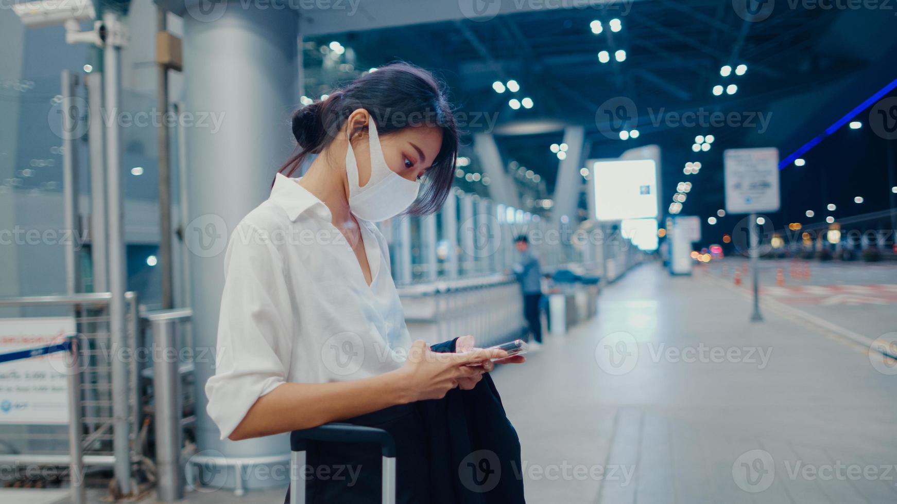 asiatisk affärsflicka anländer destination bär ansiktsmask stå utanför titta smart telefon vänta bilterminal på inrikes flygplats. affärspendlare covid -pandemi, affärsresor social distanseringskoncept. foto