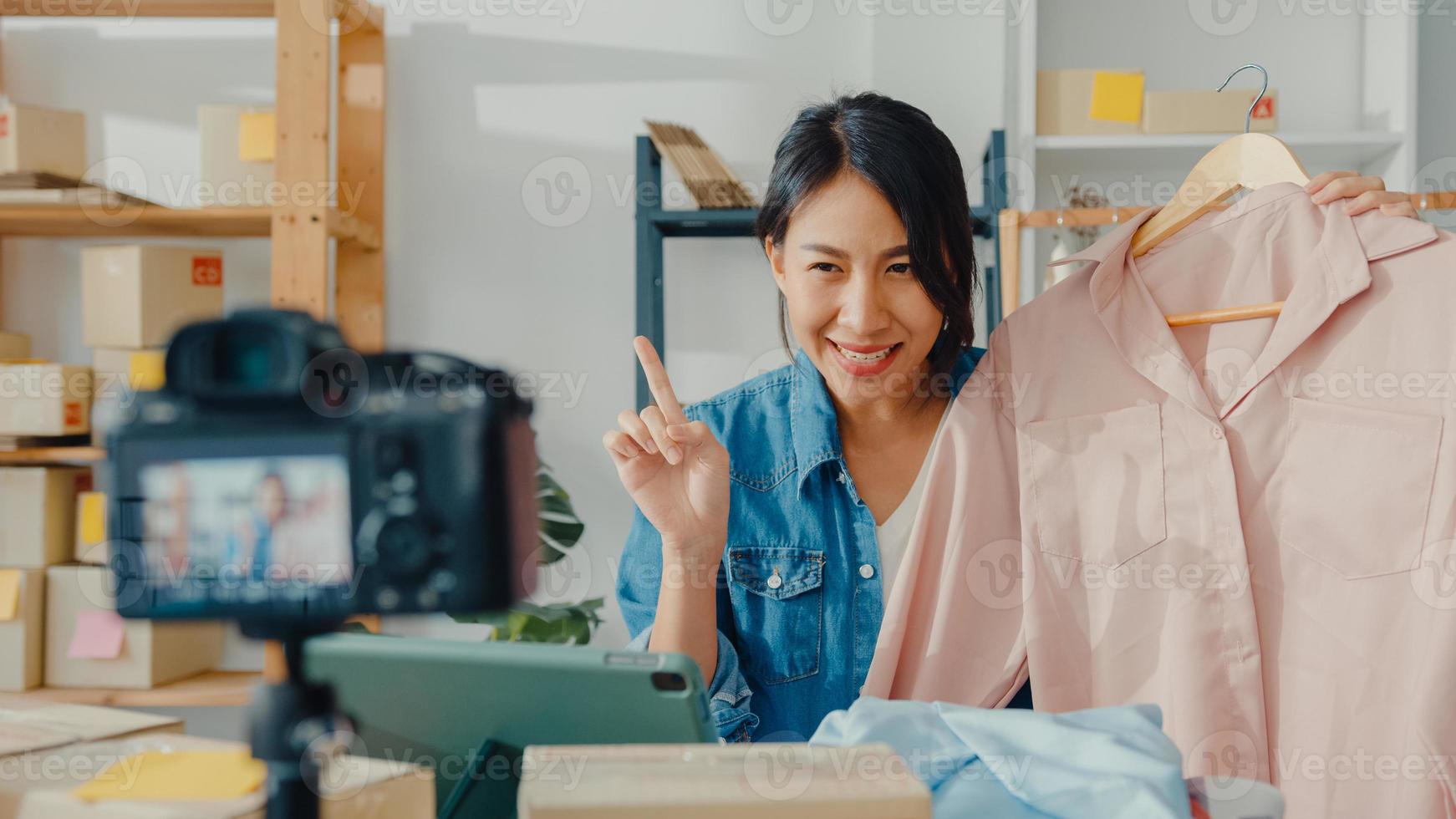 ung asiatisk dammodedesigner som använder mobiltelefon tar emot inköpsorder och visar kläder som spelar in video live streaming online med kamera. småföretagare, online marknadsföringskoncept. foto