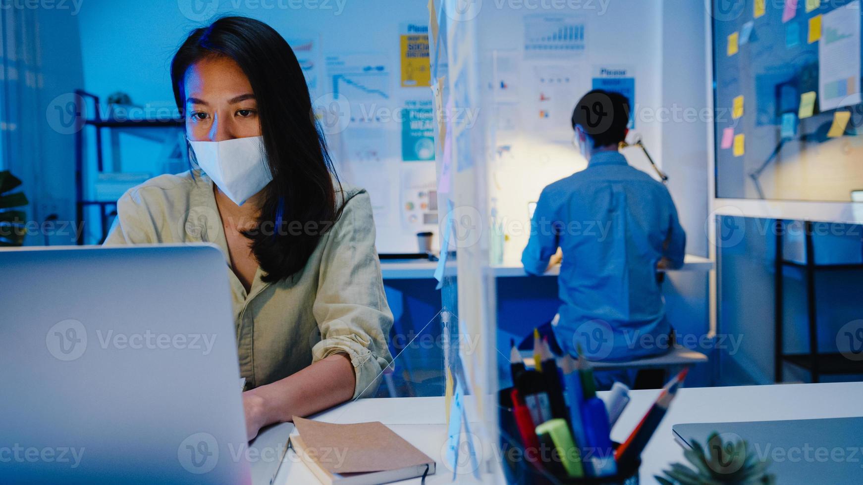 glad asiatisk affärskvinna som bär medicinsk ansiktsmask för social distansering i en ny normal situation för att förebygga virus medan du använder en bärbar dator tillbaka på jobbet på kontorsnatten. liv och arbete efter coronaviruset. foto