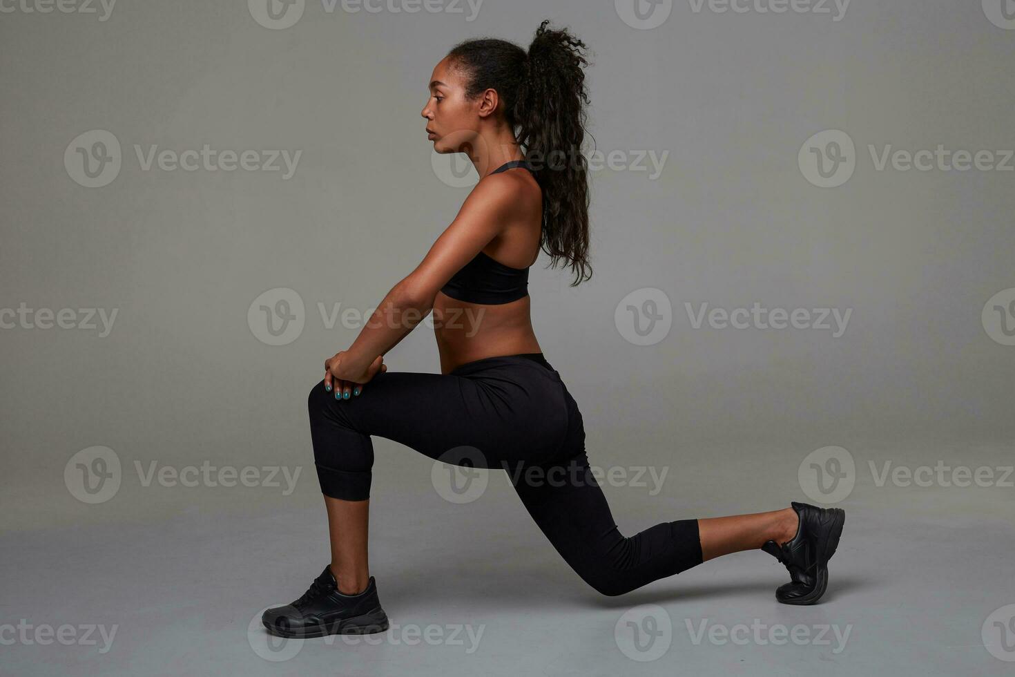 inomhus- Foto av smal Söt ung mörk flådd lång håriga brunett sports framställning stretching övningar medan Framställ över grå bakgrund i atletisk svart ha på sig