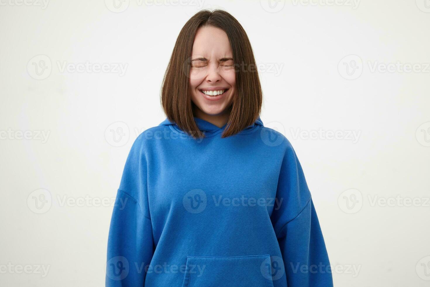 Bra ser ung Söt kort håriga brunett kvinna klädd i blå luvtröja leende glatt med stängd ögon medan stående över vit bakgrund foto