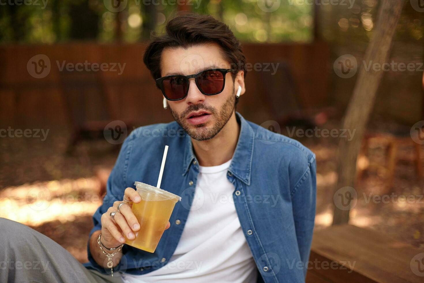 skön ung skäggig manlig ser till kamera med allvarlig ansikte, dricka juice medan Sammanträde utanför i offentlig plats, bär tillfällig kläder foto