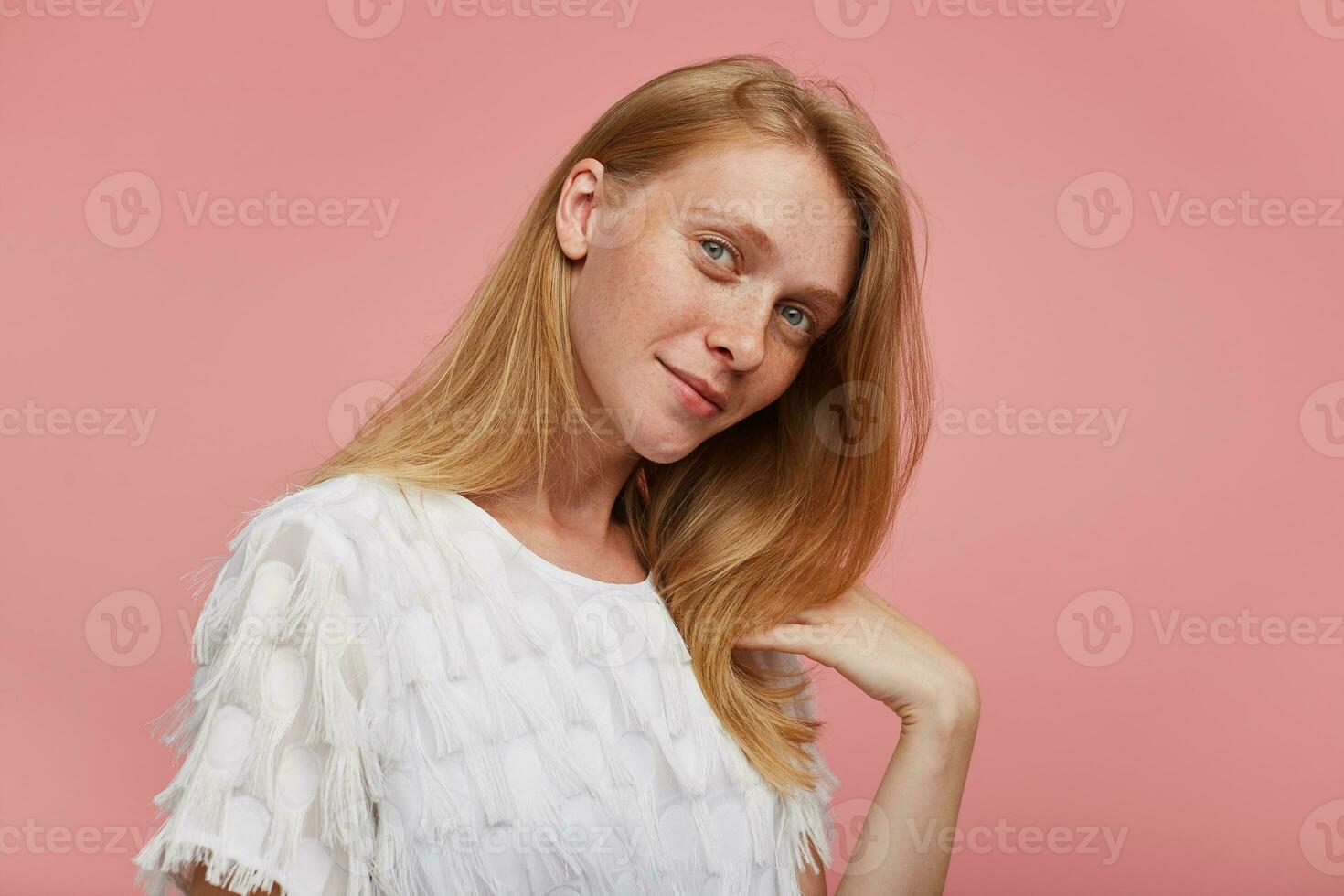 inomhus- Foto av charmig ung rödhårig kvinna med naturlig smink klädd i elegant ha på sig ser positivt på kamera och leende försiktigt, isolerat över rosa bakgrund
