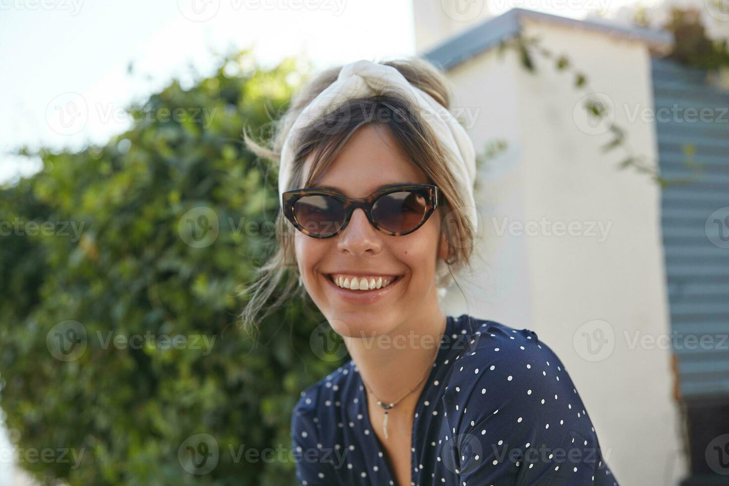 utomhus- porträtt av charmig ung brunett kvinna i årgång solglasögon bär romantisk klänning, har trevlig dag och som visar henne trevlig känslor med uppriktig leende foto