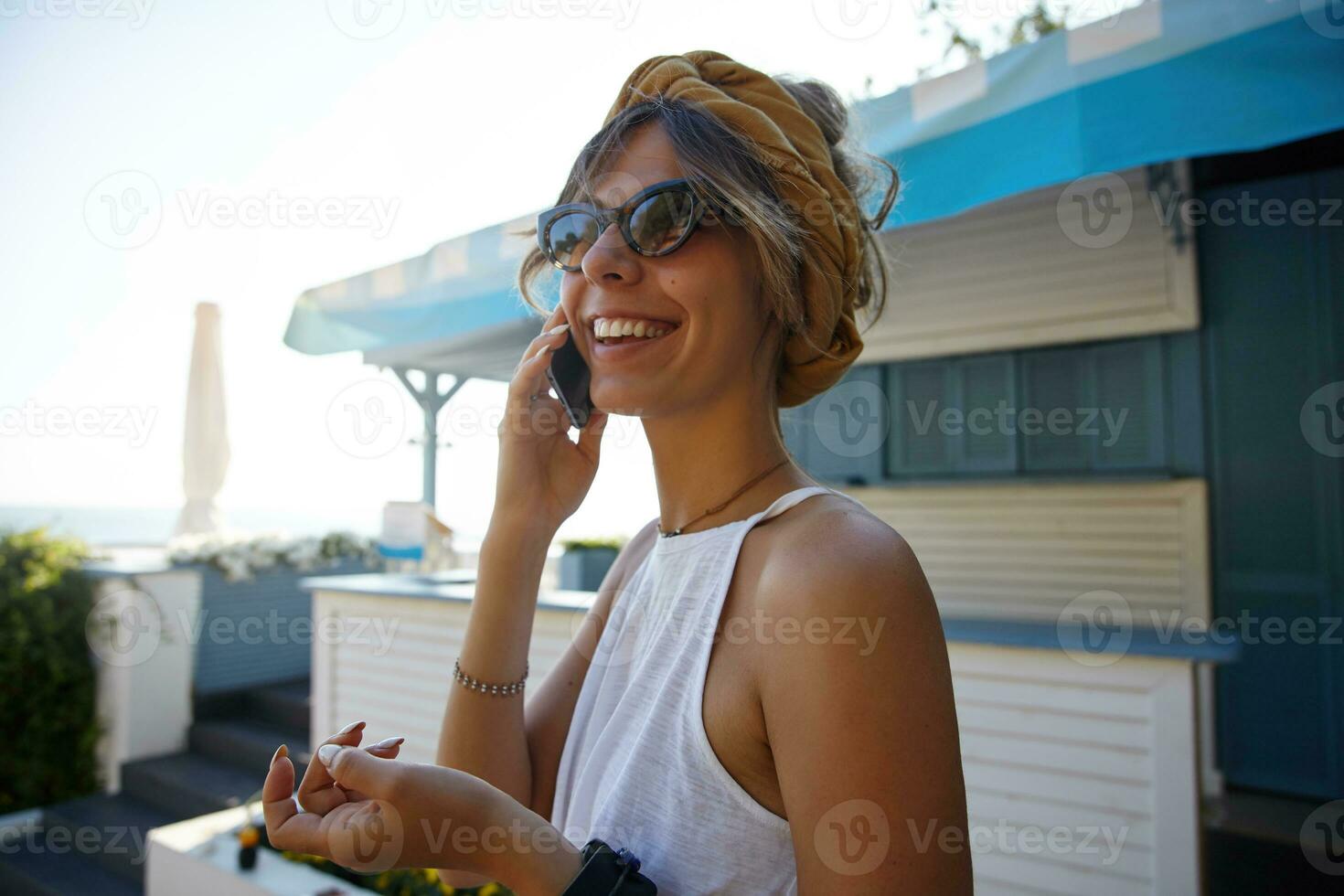 närbild av charmig ung lady i årgång trendig solglasögon bär tillfällig kläder och pannband, talande på telefon och leende allmänt medan Framställ över sommar Kafé interiör foto