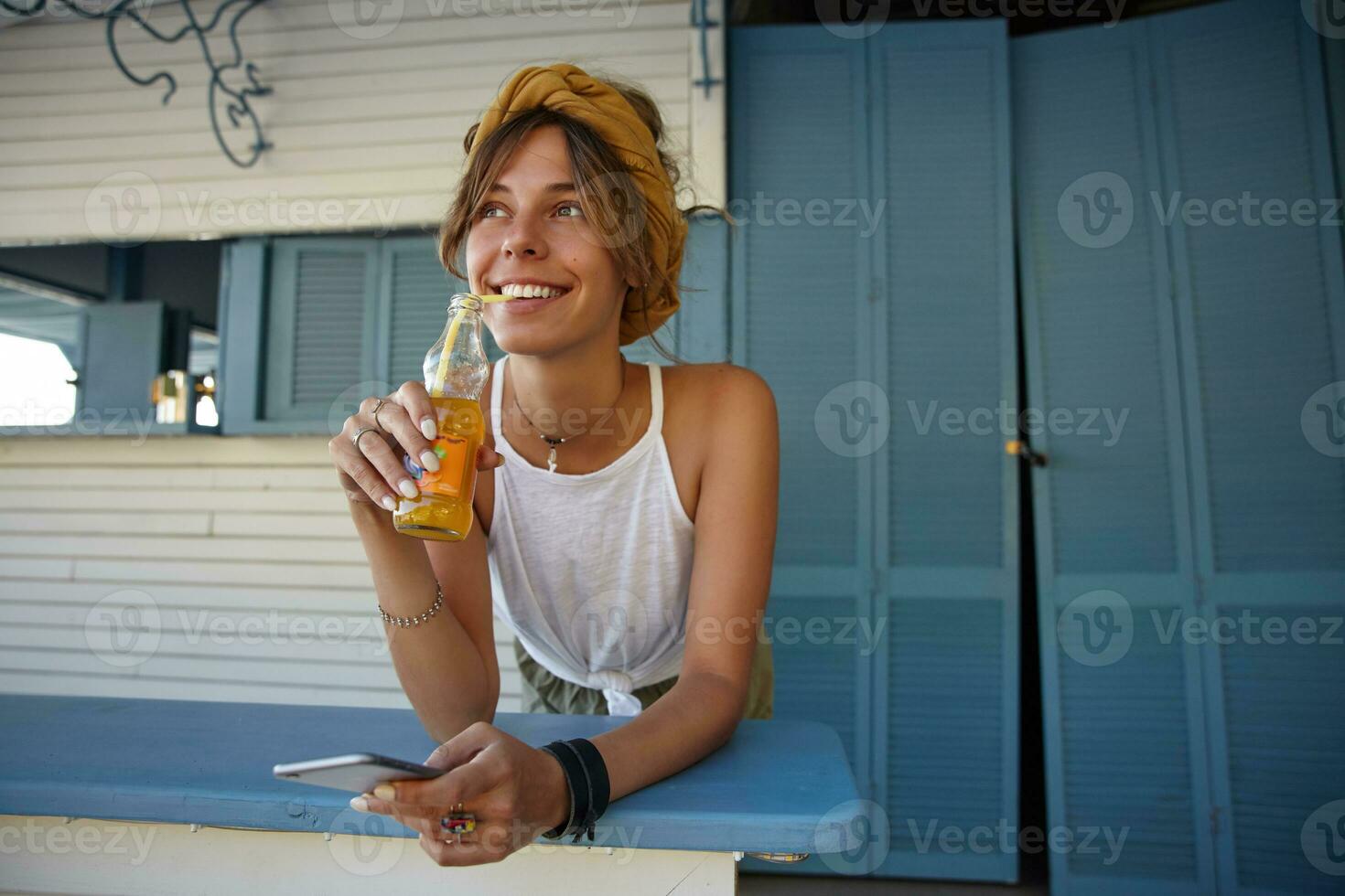 glad ung brunett lady med tillfällig frisyr bär vit topp och senap pannband och dricka citronsaft med sugrör, ser ett huvud drömmande medan stående över Kafé interiör foto