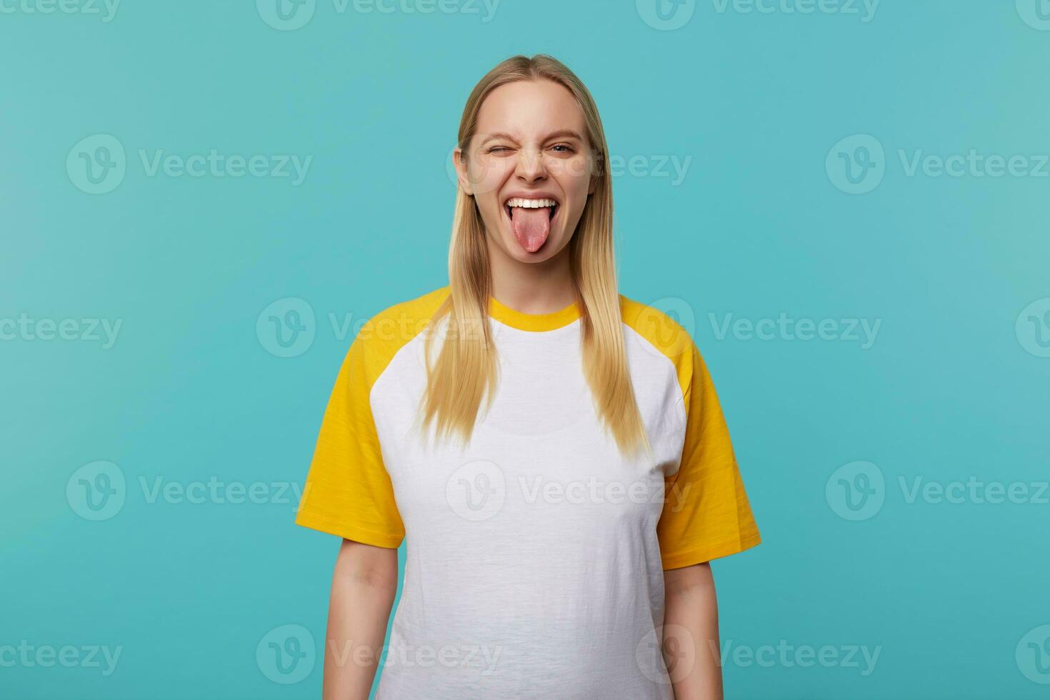 inomhus- Foto av ung Söt lång håriga blond kvinna som visar lyckligt tunga medan framställning roligt, klädd i vit och gul t-shirt medan Framställ över blå bakgrund