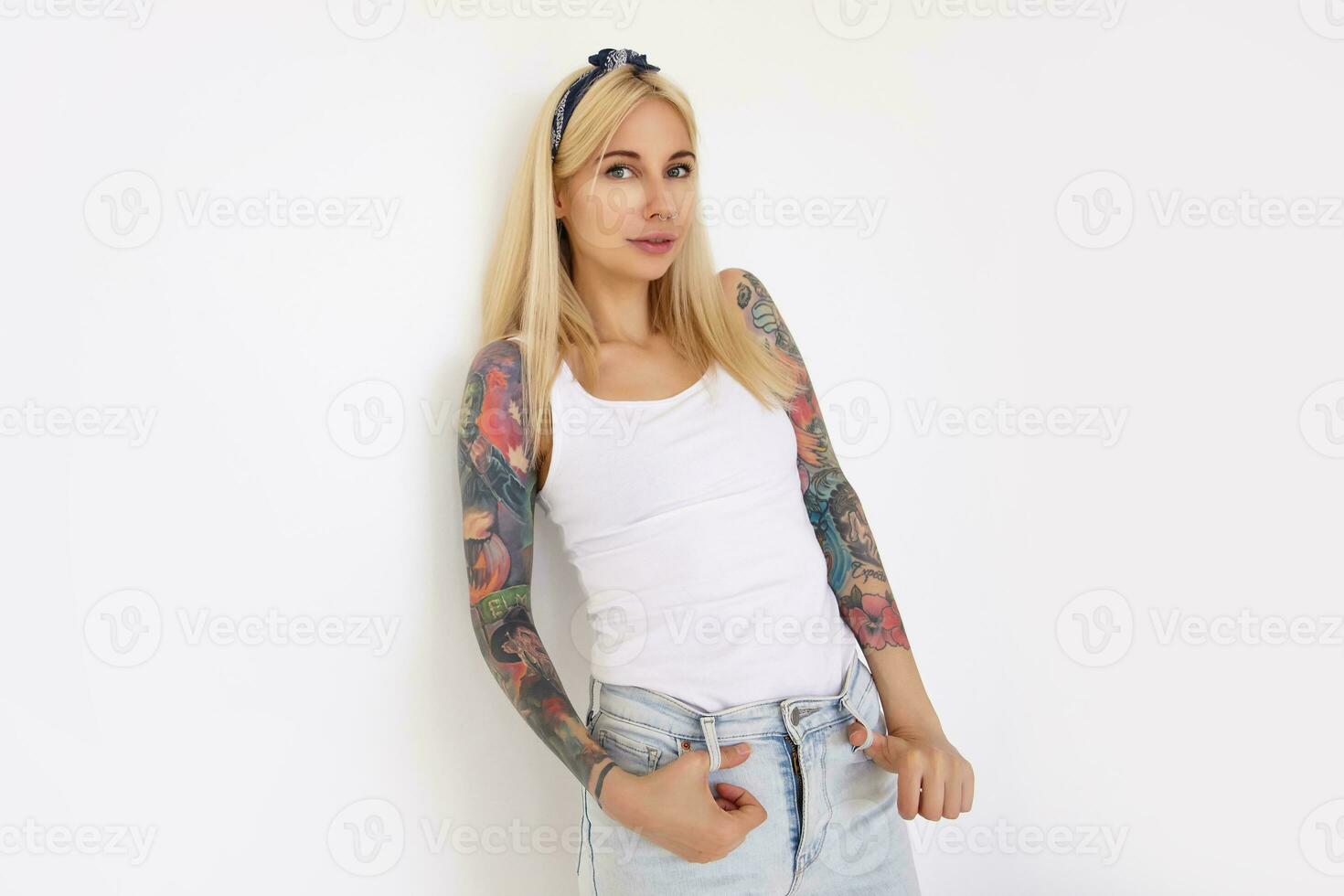 skön ung tatuerade kvinna med lång blond hår lutande henne tillbaka på vägg medan Framställ över vit bakgrund i skjorta och jeans, leende lite medan ser på kamera foto