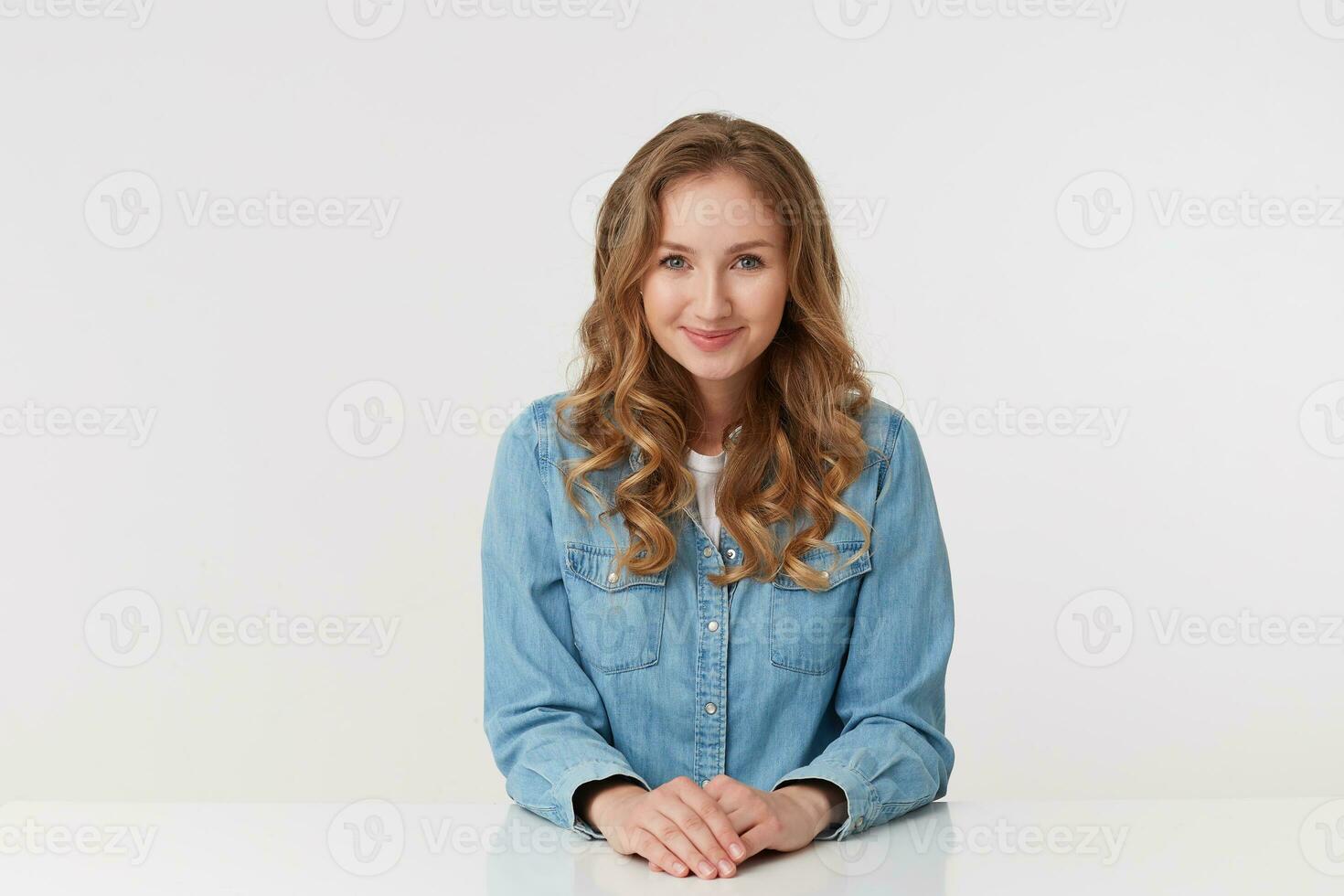 Foto av ung Söt leende ung kvinna med lång blond vågig hår, Sammanträde på de tabell med vikta vapen, bär en denim skjorta. ser på de kamera isolerat över vit bakgrund.
