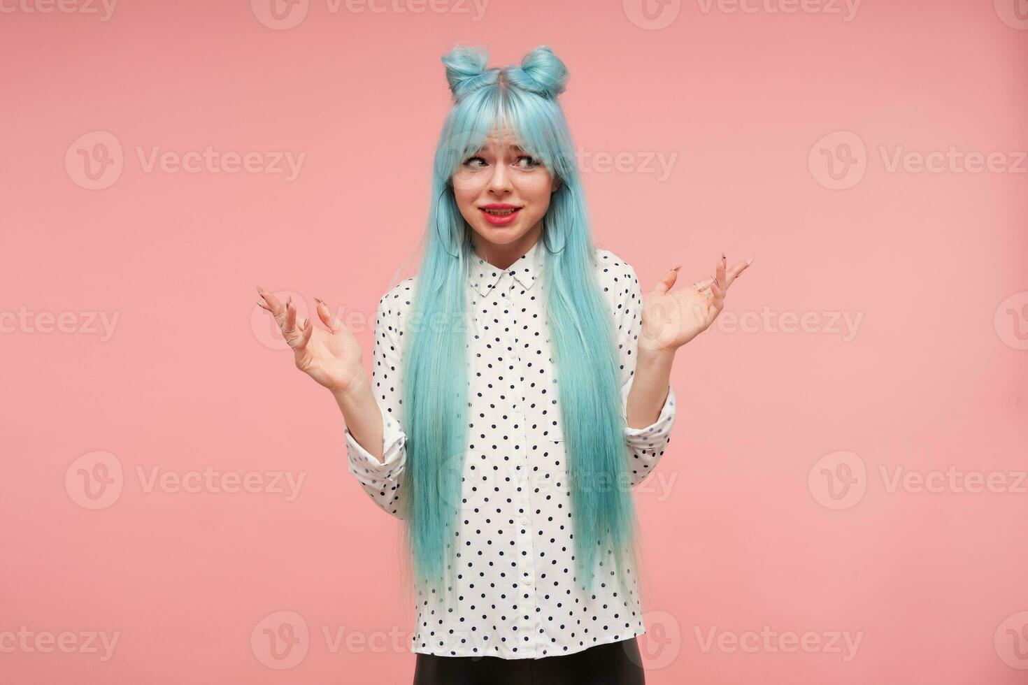 förbryllad attraktiv ung kvinna med blå anime hår höjning palmer och ser förvirrat åt sidan, skrynklig henne panna medan stående över rosa bakgrund, klädd i formell kläder foto