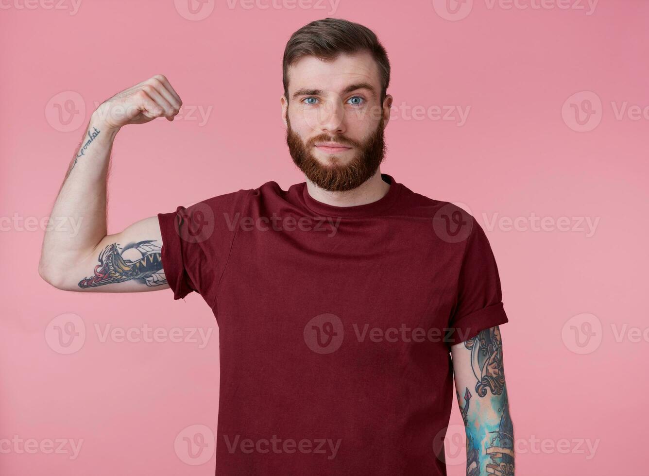 ung stilig röd skäggig man i tom t-shirt, står över rosa bakgrund, utseende Häftigt, njut av han själv och demonstrera kraftfull, utseende på de kamera och leende foto